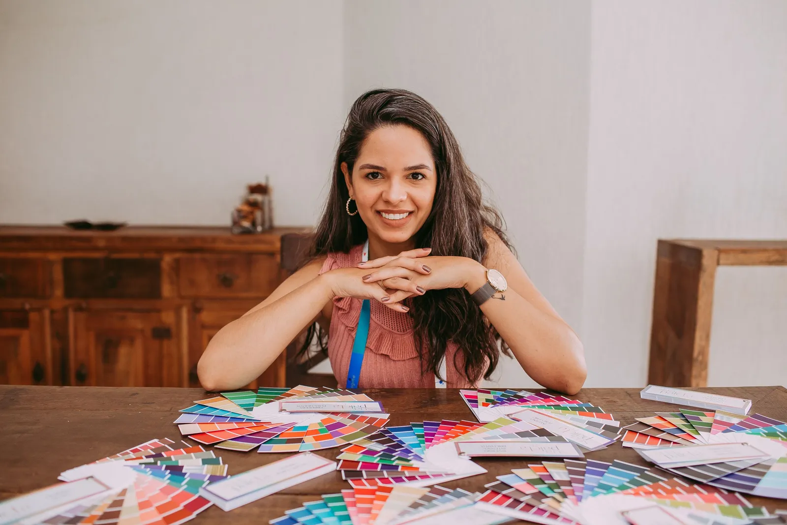 Une femme souriante est assise à une table en bois couverte de nuanciers de couleurs ouverts en éventail.