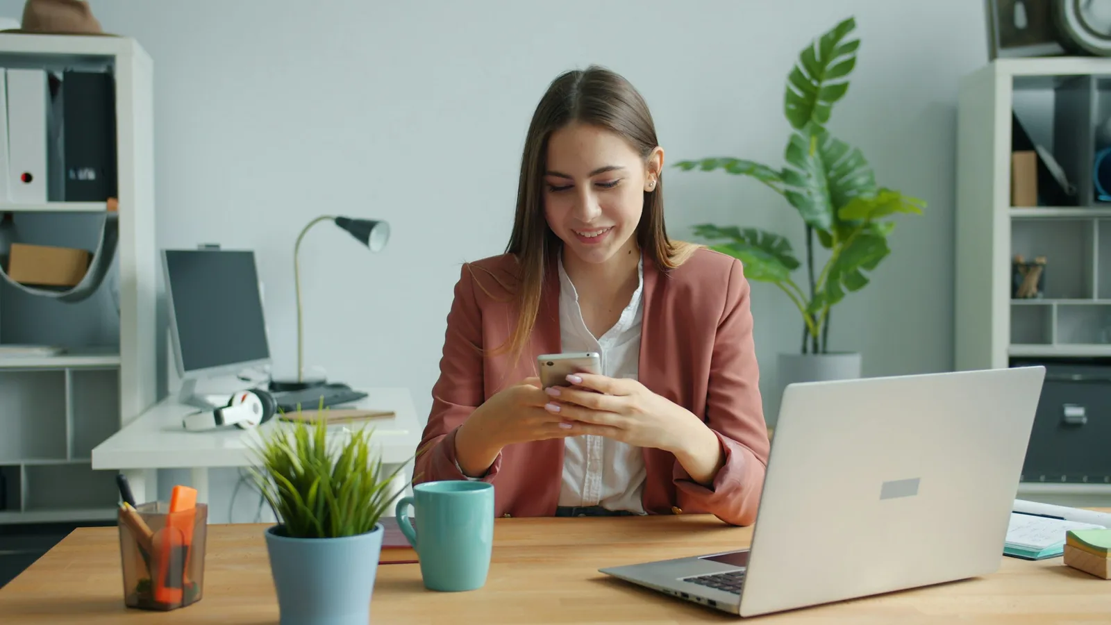 Eine junge Frau im Blazer schaut an einem Schreibtisch vor einem Laptop auf ihr Smartphone.